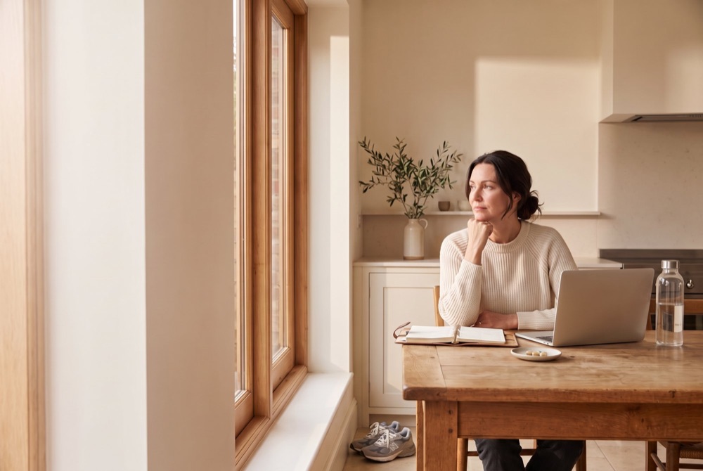 A woman at her kitchen table with a notebook and laptop, looking out of the window